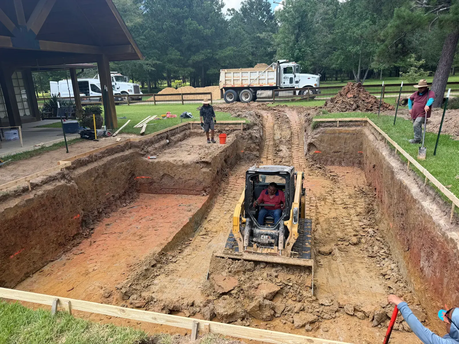 A deep pool excavation in progress with a track loader in the pit and a dump truck hauling soil on a Houston pool build